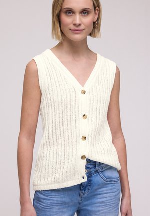 Donna che indossa un gilet lavorato a maglia color crema senza maniche, con bottoni, abbinato a jeans a vita alta di colore azzurro chiaro su uno sfondo neutro.