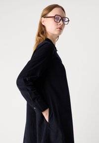 Una giovane donna con capelli lunghi, che indossa grandi occhiali geometrici e un abito scuro a maniche lunghe, in piedi di lato con una mano in tasca.