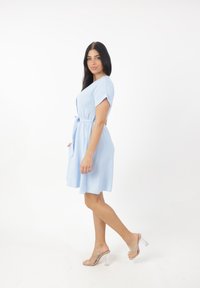 Abito azzurro chiaro a maniche corte con vita legata, realizzato in tessuto testurizzato, con gonna svasata e tasche laterali. Sandali con tacco trasparente.