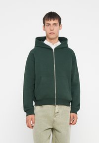 Zelena zip-up kapa iz mešanice bombaža, z kenguru žepom in rebrastimi manšetami. Nosi se čez beli srajco s ovratnikom.