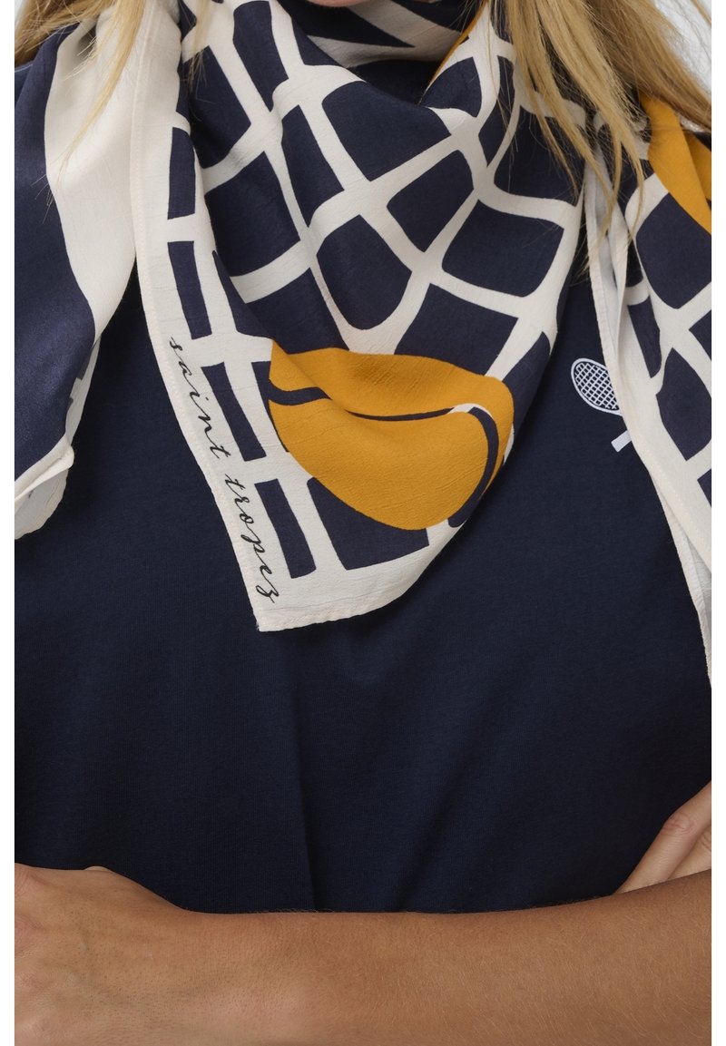 Femme portant une chemise bleu marine avec un logo de raquette de tennis, et une écharpe graphique bleu marine, blanche et moutarde avec le texte « saint tropez ».