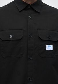 Άτομο που φοράει μαύρο πουκάμισο με κουμπιά και δύο τσέπες στο στήθος, η μία από τις οποίες έχει λευκή και μπλε ετικέτα Hugo, σε απλό φόντο.
