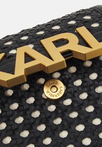 Primer plano de un bolso de mano negro tejido con grandes letras doradas que dicen "KARL" y un botón dorado redondo grabado con "KARL LAGERFELD."