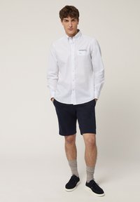 Chemise blanche à boutons avec un détail brodé bleu, associée à un short navy. Le mannequin se tient les mains dans les poches, portant des chaussettes grises et des chaussures noires.