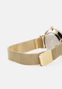 Montre avec un bracelet en mailles dorées, dotée d'un boîtier rond et d'une finition métallique lisse. Dispose d'une fermeture à boucle et d'un dos argenté avec un marquage.