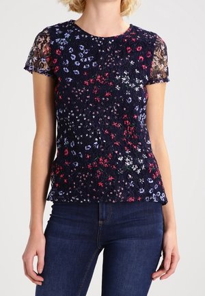 Mujer vistiendo una blusa de encaje floral negra de manga corta con flores rojas, moradas y blancas, combinada con vaqueros azul oscuro.