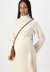 Rochie din tricot ribflat de culoare crem, cu guler înalt, mâneci lungi și talie legată. O geantă maro tip crossbody completează ținuta.
