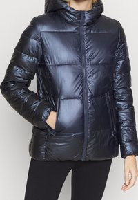 Person som bär en glänsande svart puffjacka med dragkedja fram och fickor, en hand inne i en ficka, i kombination med svarta leggings.