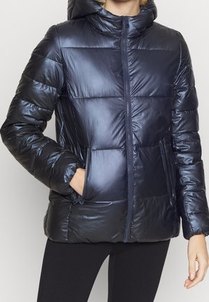 Persoon draagt een glanzende zwarte pufferjack met een ritssluiting aan de voorkant en zakken, met één hand in een zak, gecombineerd met zwarte leggings.