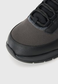 Gros plan sur l'avant d'une sneaker noire et gris foncé avec une tige en tissu, une semelle en caoutchouc renforcée et des lacets noirs sur fond clair.