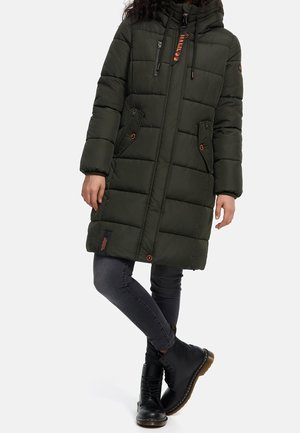 Personne portant un long manteau matelassé vert foncé avec capuche, un jean skinny foncé et des bottes noires à lacets, debout jambes croisées.