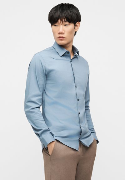 Άνδρας που φοράει εφαρμοστό ανοιχτό μπλε πουκάμισο με κουμπιά και μπεζ παντελόνια, με τα χέρια στις τσέπες, κοιτάζοντας πλάγια σε απλό φόντο.