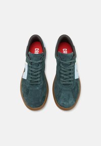 Sneaker in suede verde con suole in gomma marrone, caratterizzate da dettagli bianchi, punta tonda e design con lacci. Foderine rosse con logo del marchio ben visibile.