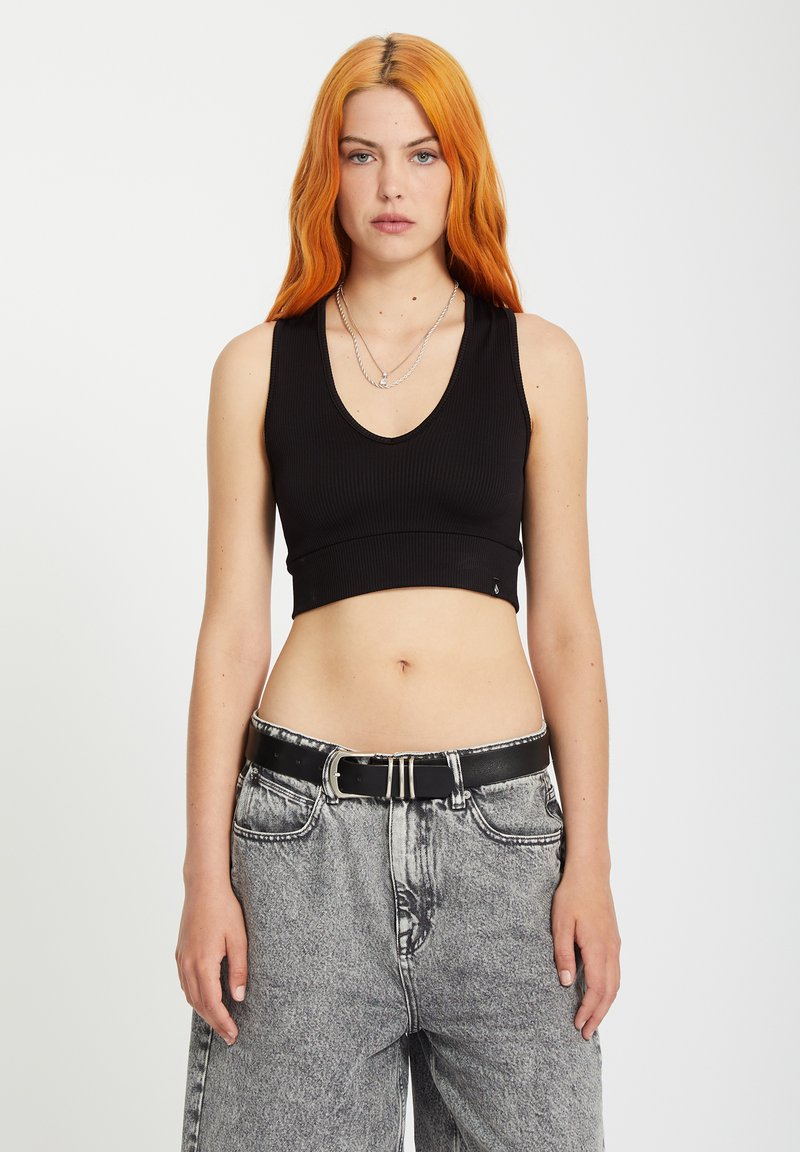 Jeune femme aux longs cheveux orange portant un haut noir court sans manches, des colliers argentés, une ceinture noire et un jean gris taille haute.