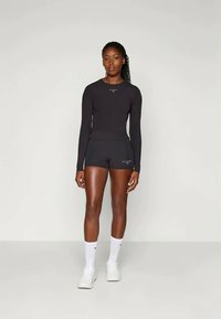 Γυναίκα στέκεται μπροστά σε απλό φόντο φορώντας μαύρο μακρυμάνικο μπλουζάκι, μαύρες βερμούδες, λευκές κάλτσες και λευκά αθλητικά παπούτσια.