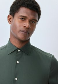 Olivgrünes Button-up-Hemd aus glattem Stoff, mit klassischem Kragen und weißen Knöpfen, taillierter Passform und klaren Linien.