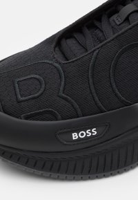 Zapato deportivo negro con parte superior de tela texturizada, detalle del logotipo en relieve y suela de goma gruesa con estrías de tracción.