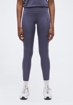 Persoon draagt donkergrijze aansluitende leggings met Puma-logo en witte sneakers met witte sokken, staand tegen een effen achtergrond.