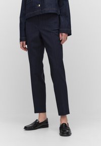 Marineblauwe pinstripe broek met een slim fit en hoge taille, gecombineerd met zwarte loafers, tegen een lichtgrijze achtergrond.