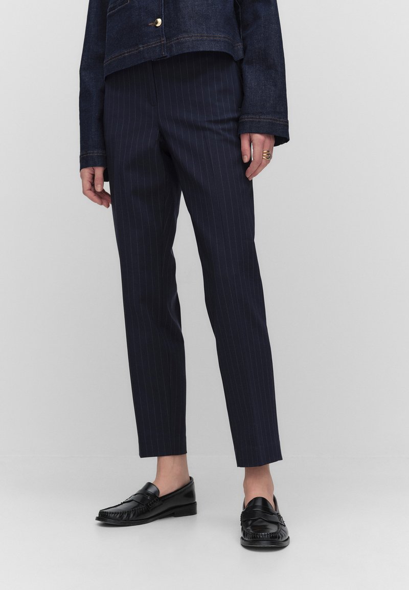 Marineblauwe pinstripe broek met een slim fit en hoge taille, gecombineerd met zwarte loafers, tegen een lichtgrijze achtergrond.