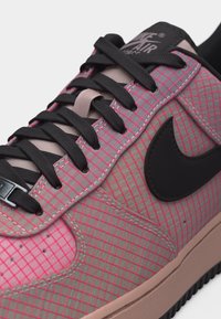 Közeli kép egy Nike Air Force sportcipőről, rózsaszín rácsmintával, fekete fűzőkkel és fekete Nike jelzéssel bézs talpon.