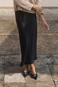 Gonna maxi nera abbinata a un maglione in maglia beige testurizzato e scarpe nere con punta decorata da fibbie, in piedi su pavimentazione in pietra.