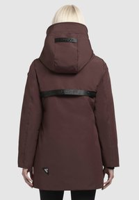 Brūns parka jakas ar augstu apkakli, regulējamu kapuci un melnu logo siksnu aizmugurē. Iezīmētais audums un zīmola emblēma.