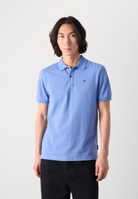 Tricou polo bleu deschis din bumbac, cu guler clasic, mâneci scurte și un mic logo brodat pe piept. Puratat cu pantaloni închiși la culoare.