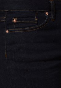 Jeans de mezclilla azul oscuro con remaches de cobre, con costuras doradas, un bolsillo frontal y una textura suave. Se puede apreciar el detalle del cinturón redondeado.