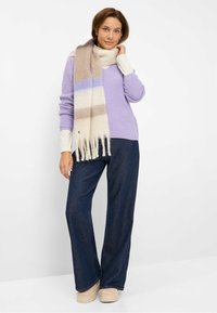 Lila gerippter Pullover, cremefarbiges langärmeliges Shirt darunter, beiger karierteter Schal mit Fransen, dunkelblaue weite Jeanshose, beigefarbene klobige Schuhe.