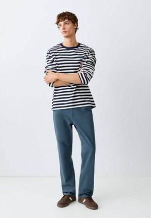 Junger Mann mit verschränkten Armen, der ein marineblau gestreiftes Langarmshirt, blaue Hose und braune Sneaker auf weißem einfarbigem Hintergrund trägt.