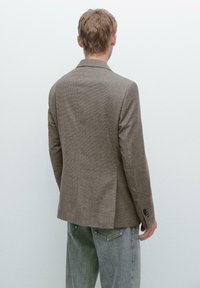 Brown check-pattern blazer with a tailored fit, notched lapels, and two back vents, paired with light gray denim jeans.