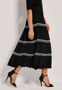 Schwarzer, gestufter Rock mit weißer, gewellter Borte, aus leichtem Stoff gefertigt. Getragen mit schwarzen, hochbelligen Schuhen mit Knöchelriemen.