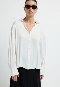 Chemise blanche à boutons avec des manches longues et une coupe décontractée. Dotée d'un col, d'une texture lisse et d'un éclat subtil. Associée à une jupe plissée noire.