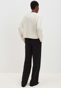 Person von hinten, die einen cremefarbenen Pullover und schwarze weit geschnittene Hosen trägt, stehend auf einem weißen strukturierten Boden vor einem schlichten Hintergrund.