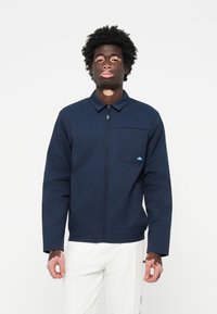 Chaqueta de navy con cremallera y cuello, un bolsillo en el pecho y un pequeño logo azul. La tela parece suave, ideal para uso casual.