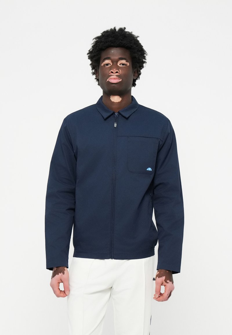 Chaqueta de navy con cremallera y cuello, un bolsillo en el pecho y un pequeño logo azul. La tela parece suave, ideal para uso casual.