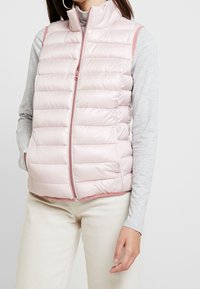 Hellrosa gesteppte Steppweste mit hohem Kragen und Frontreißverschluss. Verfügt über eine glatte Textur und Seitentaschen. Gestylt über einem grauen Langarmshirt.