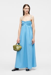 Γυναίκα που φοράει ένα μακρύ ουράνιο μπλε φόρεμα με λεπτές τιράντες, κρατάει μια μικρή τσάντα ελιάς και φοράει μαύρες μπαλαρίνες με ασημένιες λεπτομέρειες.