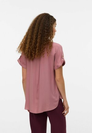 Kobieta z długimi kręconymi włosami, ubrana w luźną mauve bluzkę z krótkim rękawem i ciemnofioletowe spodnie, stojąca tyłem na jednolitym tle.