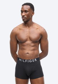 Μυώδης άνδρας στέκεται με τα χέρια πίσω από την πλάτη, φοράει μαύρα boxer με τη λέξη "HILFIGER" στη ζώνη μέσης, κοιτάζοντας προς τα αριστερά του.