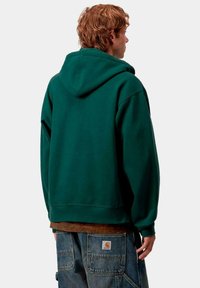 Sudadera verde con capucha de corte relajado, textura suave, bolsillo canguro y puños acanalados, combinada con pantalones cargo de mezclilla azules.