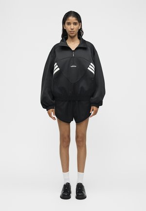 Donna in piedi rivolta verso l’obiettivo, indossa un pullover nero Adidas con mezza cerniera e strisce bianche, pantaloncini neri, calzini bianchi e scarpe da ginnastica nere.