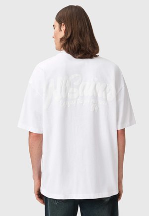 Persoon met middellang bruin haar die een los wit T-shirt draagt met vage, verhoogde tekst op de achterkant, staand tegen een effen achtergrond.