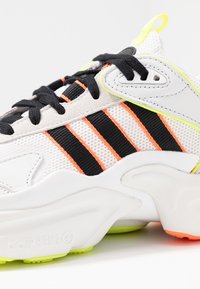 Närbild av vit sneaker med svarta och orange ränder, svarta snörningar och neongula detaljer på sulan och hälöglan.