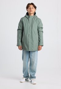Green waterproof jacket with a hood, front button closure, side pockets, and a slightly loose fit, paired with light blue jeans and white sneakers.