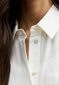 Vit linneskjorta med krage, prydd med små knappar i en ljus färg, och en bröstficka. Den synliga tygstrukturen är mjuk och något genomsiktig.
