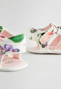 Valkoiset vauvan sandaletit, joissa on kukka- ja perhospainatuksia, vaaleanpunainen sisävuori ja säädettävä hihna, vaaleaa taustaa vasten.
