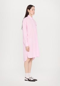 Ροζ καρό φόρεμα μακρυμάνικο με γιακά που κλείνει με κουμπιά, πλευρικές σχισμές και τσέπη στο στήθος. Μοντέλο που φοράει μαύρα και άσπρα παπούτσια.