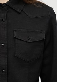 Μαύρο πουκάμισο με κουμπιά, μυτερό γιακά και μία τσέπη με καπάκι στο στήθος, φορεμένο από άτομο με μακριά μαλλιά.